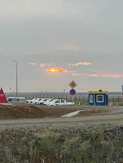 The image depicts an outdoor scene, possibly at dusk, in the vicinity of Bozshakol, Kazakhstan. The sky is overcast with some clouds tinged with orange from the setting sun. In the foreground, there is rough, unpaved terrain with some sparse vegetation. A dirt path or road runs across the middle ground.
To the left, a red triangular marker with the number "30" is visible. Behind it, a row of white vehicles, possibly utility trucks or vans, is parked. A light pole stands nearby. Further to the right, a small blue and yellow guard booth or utility shed is present. Road signs are visible, including a diamond-shaped sign with a yellow center and a circular sign with a red cross within a red circle. A chain-link fence stretches across the background, separating the area with the vehicles and shed from a distant, flat landscape. The overall impression is of an industrial or operational area, possibly related to mining or infrastructure development, given the proximity to Bozshakol, which is known for its mining operations. There are no people visible in the image.