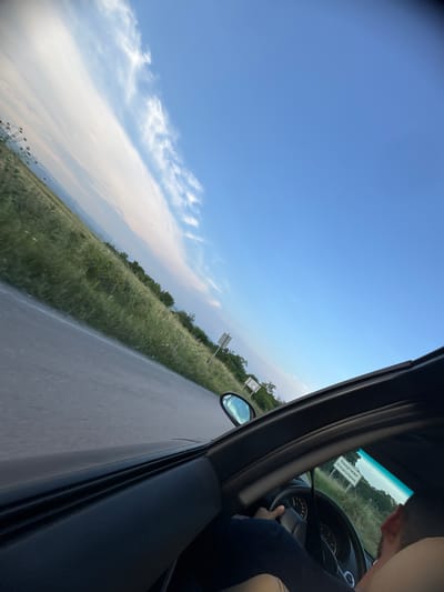 The image depicts a tilted, eye-level view from inside a moving vehicle on a road near Omarchevo, Bulgaria. The driver's arm and part of their head are visible on the lower right, with hands gripping the steering wheel. The car's black dashboard and the top edge of the open window frame the lower and right sides of the image. Through the window, an asphalt road with a faint white line on its edge recedes into the distance, bordered by a wide expanse of green grass and scattered low vegetation. Further back, a line of trees and distant rolling hills are visible. The sky occupies the upper portion, exhibiting a clear blue with streaks of white, wispy clouds stretching across the frame. A white rectangular road sign, bearing the text "Омарчево Omarchevo" in both Cyrillic and Latin scripts, is clearly visible on the right side of the road, slightly ahead of the vehicle. The driver's side mirror is also visible, reflecting a distorted portion of the sky and the road. The lighting suggests daytime, possibly late afternoon, casting an even illumination over the scene.