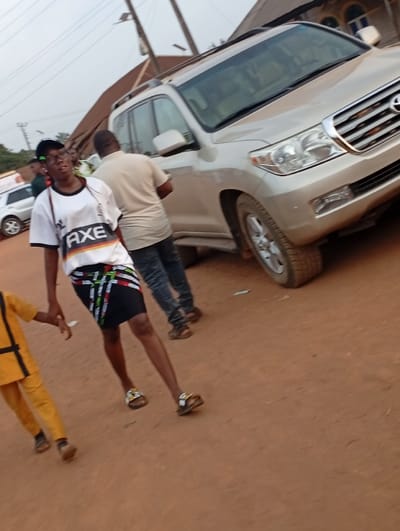 A beige Toyota Land Cruiser dominates the frame on a reddish dirt road. A person in a white t-shirt with "AXE" printed on it and a patterned skirt walks in front of the vehicle. Behind them, another individual in a tan shirt and jeans stands beside the car. A child in a yellow outfit partially enters the frame from the left. Overhead wires and a portion of a building are visible in the background.