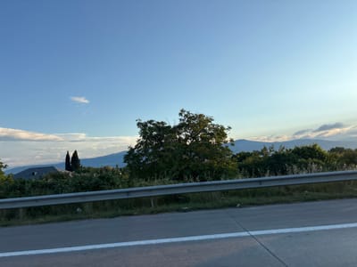 The image captures a scenic view of a rural landscape in Gorovani, Georgia, likely taken from a moving vehicle on a sunny day. A paved road with a white dividing line runs across the foreground, indicating it is a highway or main road. A silver guardrail separates the road from the embankment. Beyond the guardrail, a lush green area is visible, dotted with trees and bushes. In the mid-ground, a large, leafy tree stands prominently, with a cluster of dark green, conical trees and a roof of a building in the background. Further in the distance, rolling hills and mountains are silhouetted against a clear blue sky with a few wispy clouds. The overall impression is one of natural beauty and tranquility, characteristic of the Georgian countryside. There are no people or animals visible in the image.