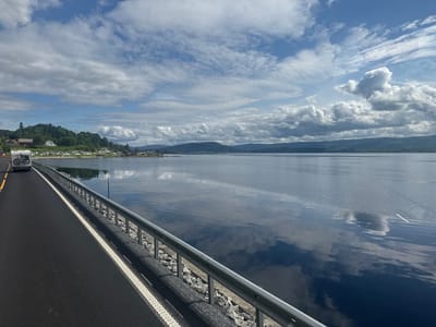 The image is a wide-angle, eye-level outdoor photograph taken from a road, likely a bridge, overlooking a large body of water. The road surface is dark asphalt with white and yellow lane markings. A metal guardrail runs along the edge of the road, separating it from the water. Small, gray rocks are visible beneath the guardrail.
On the left side of the road, a white recreational vehicle (RV) with bicycles mounted on the rear is traveling away from the camera. In the distance, across the water, a tree-covered hillside is visible, with several buildings and what appears to be a small village or settlement nestled on the shore. The sky is partially cloudy, with dramatic formations of white and gray clouds against a bright blue sky. The water is calm and reflects the sky and clouds.
Based on the description of the location as Snåsa, Norway, this scene likely depicts a view of Snåsavatnet, one of the largest lakes in Norway, with the surrounding landscape typical of the region. The RV suggests a person or people are traveling, likely on vacation or a road trip. The overall impression is one of natural beauty and tranquility.