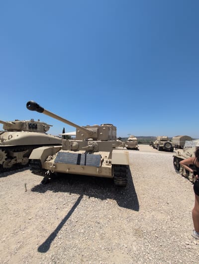 In an outdoor museum setting, several tan-colored military tanks are displayed on a gravelly ground under a clear blue sky. The foreground features the front half of a tank with a long cannon barrel. To its left, another tank, possibly a Sherman, is partially visible. In the background, a line of more tanks and military vehicles extends towards a distant, hazy landscape with rolling hills and some greenery. A person's legs and lower torso, wearing shorts and a tank top, are visible on the far right, facing away from the camera and towards the tanks. The overall scene depicts a sunny day at a historical military vehicle exhibition.