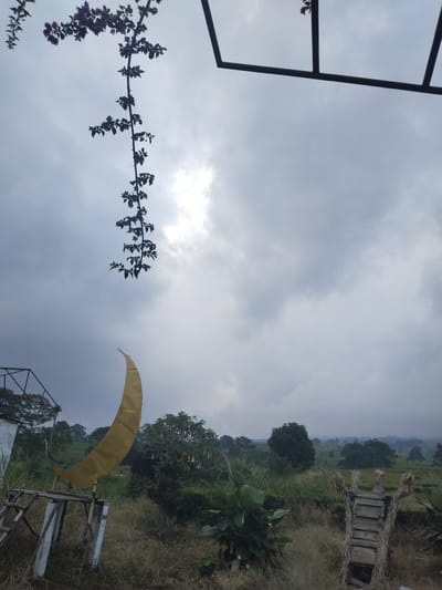 This image depicts an outdoor scene likely in Pagar Alam, Indonesia, under an overcast sky. In the foreground, a large, golden crescent moon sculpture is mounted on a rustic wooden structure. To the left, part of a white building and a metal frame are visible. A vine with purple flowers hangs down from the upper left corner. The middle ground features dry grass, bushes, and trees, with rolling hills and more vegetation in the background, suggesting a rural or natural landscape. A wooden walkway decorated with straw is situated in the lower right portion of the image. The overall atmosphere is subdued due to the cloudy weather. There are no people or animals visible in the frame.