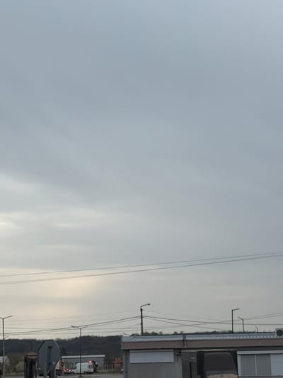 The image depicts a sky with overcast, grey clouds. The lower right side of the frame features power lines, utility poles, and parts of buildings. It's a view in Plodovitovo, Bulgaria. A treeline and parts of vehicles are visible in the distance, partially obscured by the utility infrastructure.