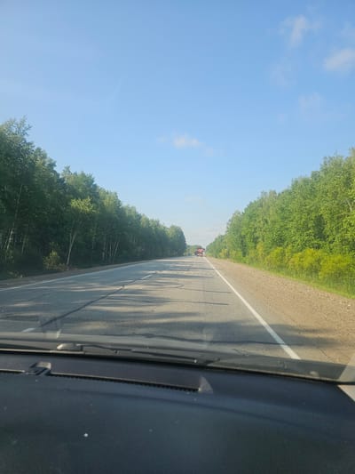 The image shows a view from inside a vehicle driving on a paved road lined with trees. The road has lane markings and appears to be a highway. In the distance, there are two trucks on the road. The sky is clear blue with a few wispy clouds. The environment appears to be a rural or suburban area, possibly in the city of Zoevka, Russia, given the provided context. The image is taken during daylight hours, and the lighting suggests it could be morning or late afternoon. There are no visible people in the image, and the focus is on the road and the surrounding landscape. The interior of the vehicle, including the dashboard and windshield wipers, is visible in the foreground, indicating the perspective is from the driver's or passenger's seat.