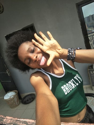 The image displays a close-up selfie of a young, dark-skinned individual with short, textured black hair, consistent with an afro hairstyle. The subject is positioned facing slightly right, with eyes closed and tongue extended playfully. The right hand is raised, fingers splayed, partially obscuring the upper left side of the face. The individual wears a dark green sleeveless crop top with white trim, featuring "BROOKLYN" in white capital letters across the chest. Multiple dark, beaded bracelets adorn the left wrist. A navel piercing is visible on the exposed abdomen.

The background indicates an outdoor urban environment, consistent with a balcony, porch, or terrace in Cotonou, Benin. To the left, a light-colored woven plastic basket with a lid and a dark, circular object rest on a light-tiled floor. A dark door frame is visible behind these items. To the right, a window or glass panel reflects distant multi-story buildings, typical of a city landscape. The wall directly behind the subject is painted a light grey. A horizontal light-colored surface, possibly wood or painted concrete, occupies the bottom foreground. No specific street address or unique landmark is identifiable to pinpoint the exact location within Cotonou, Benin, beyond the general context of an urban outdoor residential or commercial space.