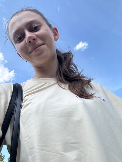 This is a low-angle, wide shot selfie taken outdoors on a bright, sunny day. The sky is a clear, vibrant blue with scattered, fluffy white clouds. A young person with brown hair is looking up at the camera, their face filling the left side of the frame. They have a light complexion and appear to be smiling faintly. Their hair is long and wavy, and some strands are blowing in the wind. They are wearing a light-colored t-shirt with some faint red text visible on the right shoulder area, though it is mostly obscured by their hair and the angle of the shot. A black strap, possibly from a bag, runs across the lower part of their torso. The perspective suggests the photo was taken looking up towards the sky, with the person's face angled down towards the camera. The overall mood is casual and relaxed, possibly indicating a pleasant day spent outdoors in Bürgstadt, Germany. The text on the t-shirt is partially visible and appears to be in a script font, but it is not legible enough to discern specific words.