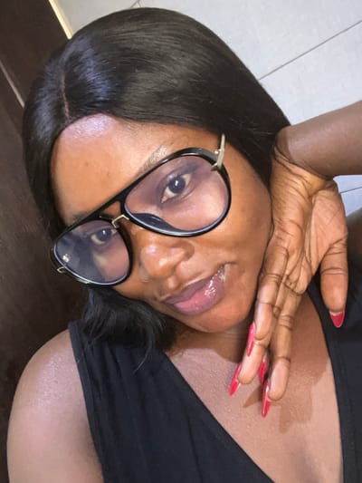 A close-up portrait features an adult female with a dark complexion, long dark hair parted at the crown, and wearing black aviator-style glasses with clear lenses. Her eyes are visible through the lenses, directed towards the viewer. Her lips are coated with a glossy, light-toned product. She is dressed in a black sleeveless garment. Her left hand is raised, with fingers splayed and resting against her right cheek/jawline. Her fingernails are long, pointed, and painted red. The background is an indistinct light-colored, possibly tiled or paneled, interior wall. This scene is located in Dzhidu, Nigeria.