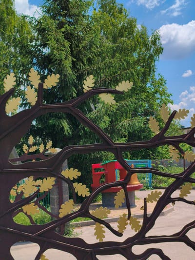 This is a medium-shot, eye-level, outdoor photograph taken on a sunny day in the city of Velyka Bahachka, Ukraine. The foreground is dominated by a dark brown, stylized metal tree sculpture with multiple branches and yellow oak leaf cutouts. The sculpture is in sharp focus, with its intricate design obscuring parts of the background.

Behind the sculpture, a playground is partially visible. A bright red, curved structure, possibly a slide or a bench, is situated on a circular concrete base. To the right of the red structure, a blue metal fence or railing is present, and further to the right, a glimpse of a building with a tiled roof can be seen.

The background is filled with lush green trees, primarily coniferous trees with dense foliage, and some deciduous trees with lighter green leaves. The sky above is a vibrant blue with scattered white, fluffy clouds. The ground in the playground area appears to be paved with light-colored rectangular stones. The overall impression is that of a park or a public recreational space.