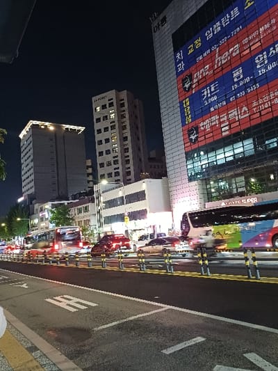 This nighttime image depicts an urban street scene in 서대문구, South Korea. The perspective is low, looking across a multi-lane road from what appears to be a curb or sidewalk. On the right, a prominent multi-story commercial building features extensive vertical illuminated signage with red and blue panels displaying Korean text, including "치과" (dentist), phone numbers such as "02-322-7291", "here", and "15.000-", alongside a stylized bull logo. The building's top edge includes the Korean text "아름" (Areum). To the center-left, a narrower high-rise building is visible with "LA FORET" at its peak, adjacent to a third, broader high-rise with an illuminated rooftop edge further to the left.
The foreground consists of dark asphalt pavement with white road markings, including a double solid line, a dashed line, and a partially visible '버스' (bus) lane symbol. A dark, circular patch is present on the asphalt. A series of yellow and black flexible bollards delineate the road lanes. Traffic is in motion, evidenced by motion blur on several vehicles: a red and white bus on the left, a multi-colored bus (blue, green, pink, yellow) with "LOTTO SUPER" text visible above it on the right, and multiple cars with illuminated headlights and taillights. Streetlights illuminate trees lining the left side of the road. The sky is dark.