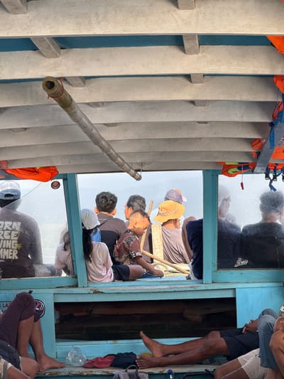 This is a photograph taken from inside a boat in Desa Pemenang Barat, Indonesia. The boat appears to be made of wood with a blue painted interior. Several people are seated inside the boat. In the foreground, a person's legs and feet are visible, along with some items on the floor including red flip-flops and a plastic bag.
Looking through a window or opening in the boat, more people are visible seated in rows. They are looking in different directions, some towards the water and some towards each other. Some of the people are wearing hats, including a child with a patterned shirt. Orange life vests are hanging from the side of the boat.
The environment outside the boat appears to be a body of water with a foggy or overcast sky. A cylindrical object, possibly a piece of bamboo, is suspended from the ceiling of the boat with rope. The overall scene depicts a group of people traveling by boat in a coastal or island setting.