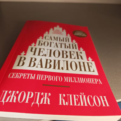 A close-up shot shows a red book titled "Самый богатый человек в Вавилоне" (The Richest Man in Babylon) by Джордж Клейсон (George Clason). The subtitle reads "Секреты первого миллионера" (Secrets of the First Millionaire). The book sits on a grey surface. The image was taken in Pokrovskoye, Russia.