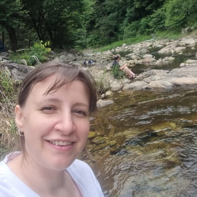 In an outdoor scene, a woman is pictured from the chest up, taking a selfie. She has fair skin, dark brown hair styled in a bob with bangs, and is wearing a white shirt and silver earrings. She is smiling broadly, showing her teeth. In the background, a shallow, rocky stream flows through a lush, wooded area. Two other individuals are visible in the background, sitting on the rocks by the stream. The environment is natural and appears to be a park or natural reserve. This image was captured in Mureungdowon-myeon, South Korea.