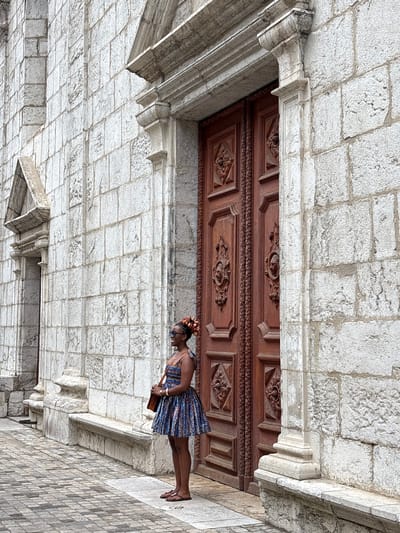 A woman stands in front of a large, ornate wooden door set within a stone building. She is wearing a short, patterned dress, sandals, and sunglasses, and is holding a brown bag. The stone building has a textured, light-colored facade. The ground is paved with cobblestones. The scene appears to be in a historic European city, likely Annecy, France, given the architectural style of the building and the cobblestone street.