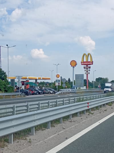 This image captures a roadside scene in Studena, Bulgaria, under a partly cloudy sky, suggesting it is daytime. The primary subjects are commercial establishments along a highway.
Prominently featured is a McDonald's restaurant with its iconic golden arches and a "McDonald's" sign. Below it, smaller signs indicate "McCafe" and "McDrive," along with the operating hours "0-24," signifying it's open all day. Adjacent to the McDonald's is a Shell gas station, identifiable by its distinctive yellow and red canopy and Shell logo. There are several cars parked at the gas station and in front of the McDonald's, suggesting customers are actively using these services. A white truck is visible on the highway, indicating ongoing traffic.
The setting is a highway with a clear lane separation, marked by a metal guardrail and white lane lines. The area around the highway includes grassy verges and scattered vegetation. In the background, trees and some buildings can be seen. The overall impression is one of a typical service area along a busy road, providing food and fuel to travelers. There are no discernible emotions conveyed in the image, as the focus is on the commercial landscape and infrastructure.