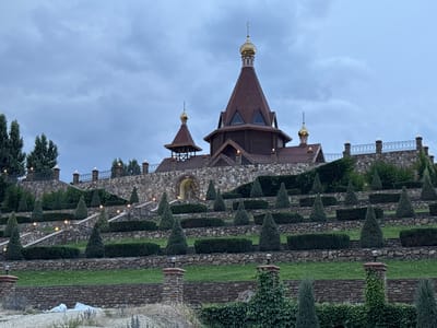 This media file depicts a beautiful, tiered garden leading up to a wooden church with golden domes. The garden features numerous stone terraces, meticulously planted with rows of manicured evergreen trees and hedges. Small, bright lights are strategically placed along the terraces and pathways, illuminating the scene, suggesting it might be dusk or evening. The church itself is made of dark wood with a prominent, steep-roofed main structure and a smaller bell tower. Both structures are crowned with golden domes topped with crosses, indicative of Orthodox Christian architecture.
The scene is set on a cloudy day, with a soft, diffused light that creates a serene and somewhat melancholic atmosphere. The layered landscaping and the imposing architecture suggest a place of religious significance or a grand estate. The use of stone and natural elements, combined with the careful cultivation of the greenery, speaks to a desire for both natural beauty and ordered design. There is no visible text in the image. The location context is Staraya Stanitsa, Russia.