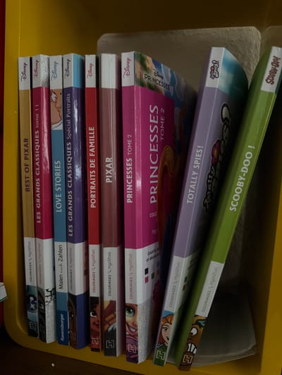 The image shows a close-up, eye-level view of several Disney-themed books stacked vertically on a yellow shelf. The books are arranged from left to right, with their spines facing the viewer. The titles visible on the spines include "BEST OF PIXAR," "LES GRANDS CLASSIQUES tome 1," "LOVE STORIES," "LES GRANDS CLASSIQUES Spécial Portraits," "PORTRAITS DE FAMILLE," "PIXAR," "PRINCESSES TOME 2," "Totally Spies!", and "SCOOBY-DOO!". Many of the books also have smaller text indicating they are coloring books with mysteries. The shelf is part of what appears to be a child's bookshelf or storage unit. The background is out of focus but suggests an indoor environment. There are no people or animals present in the image, and no specific landmarks or street names are visible, so the location within Recklinghausen, Germany, cannot be determined. The lighting is adequate, and the image is in focus, allowing for clear identification of the books and their titles.