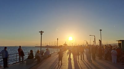 This media file depicts a picturesque sunset scene along a bustling coastal promenade, likely in Alimos, Greece. The time of day is late afternoon or early evening, with the bright, golden sun positioned low and centrally on the horizon, casting a warm glow across the entire image.
Numerous people are the primary subjects, engaged in various leisure activities. They are mostly adults, walking, strolling, standing, and sitting along the paved promenade. Many figures are silhouetted against the intense sunlight, creating long, dark shadows that stretch across the ground towards the viewer. Some individuals are seen leaning on the metal railings, looking out towards the sea, while others are seated on concrete benches. On the far left, two men are clearly visible, one in a light blue jacket and the other in a red shirt.
The setting is a waterfront area with a marina. To the left, the calm blue sea extends to the horizon, dotted with faint outlines of distant land or islands. To the right of the promenade, a dense collection of boats and yachts fills the marina, their masts creating a complex pattern of dark silhouettes against the bright sky. Several tall lampposts line the promenade, also appearing as stark silhouettes. A small, light-colored building or kiosk is visible on the far right edge of the frame.
Notable details include the clear, vibrant sky, transitioning from a deep blue at the top to fiery orange and yellow hues closer to the sun. A distinct lens flare is visible near the sun's brilliance, and a faint white contrail can be seen in the upper right quadrant of the sky. The overall atmosphere appears peaceful and communal, as people gather to enjoy the scenic sunset. No visible text is present in the image.
