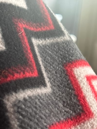 A close-up, indoor, macro photograph displays the textured weave of a blanket or fabric with a geometric zig-zag pattern in black, red, and white. The focus is on the material's surface, revealing individual fibers and the raised texture of the knit. The lighting suggests an ambient, possibly natural, light source casting soft shadows. No specific location in Richmond Hill, Canada, can be identified from the image. There are no people, actions, or interactions depicted.