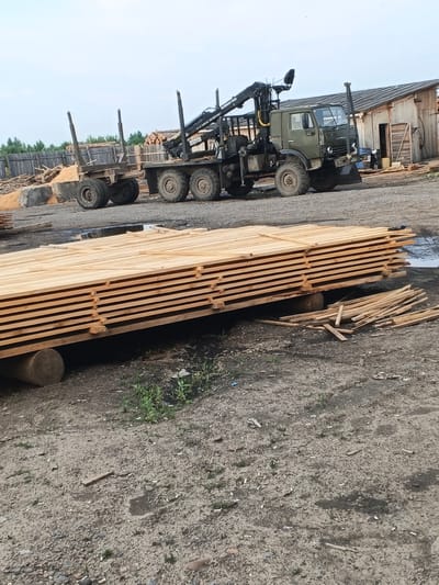 This media file depicts a lumber yard in Strelka, Russia. The primary subjects are a green military-style truck with a crane attachment and a wooden trailer loaded with logs. A large stack of lumber is prominently featured in the foreground, resting on wooden beams and a large log. In the background, a rustic wooden building, possibly a workshop or storage facility, stands to the right, with a wooden fence and more lumber visible behind it. Scattered wood scraps and debris are on the ground.
The scene suggests a logging or lumber processing operation in progress or recently completed. The truck and trailer indicate the transportation of raw materials, while the stacked lumber points to processed wood. The overall atmosphere is industrial and utilitarian. The time of day appears to be daytime, with overcast or cloudy skies, suggesting moderate weather conditions. There are no people visible in the frame, emphasizing the focus on the machinery and materials.
Notable details include the robust tires of the truck and trailer, designed for rough terrain. The crane on the truck is positioned as if it has been used to load or unload logs. The neatly stacked lumber in the foreground highlights the orderly nature of the processed material. The wooden building in the background has a weathered appearance, typical of structures in rural or industrial settings. The ground is a mix of dirt and gravel, with patches of sparse vegetation.
There is no visible text in the image that can be clearly read or identified.