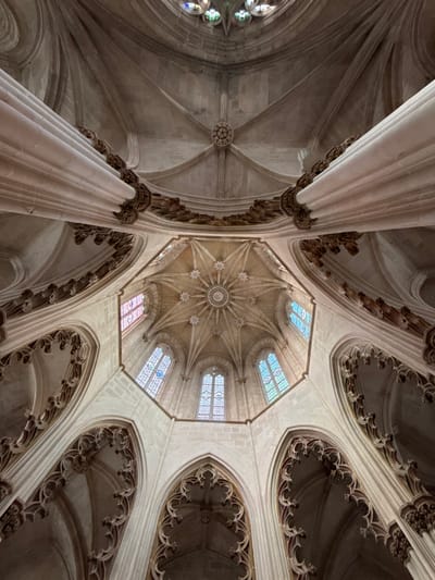 The image displays an upward-looking view into the ornate vaulted ceiling and upper walls of an octagonal chapel within the Batalha Monastery, located in Batalha, Portugal. The central feature is an elevated octagonal dome, constructed from light-colored stone, characterized by intricate ribbed vaults converging towards a prominent, circular carved boss. Smaller star-shaped bosses are present at rib intersections. Around the base of this central dome are tall, slender arched windows featuring stained glass in various hues, including blue, red, pink, and clear patterns, allowing natural light to penetrate. Supporting elements include massive, fluted, cream-colored columns with elaborately carved capitals and friezes depicting foliage motifs, which are positioned at the periphery of the central octagonal structure. Below these columns and extending downwards, pointed arches with decorative tracery are visible. The overall architectural style is consistent with late Gothic and Manueline periods. No individuals or movable objects are present in the scene.