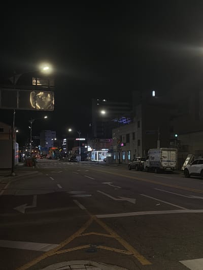 The image depicts a nighttime street scene in Dongducheon-si, South Korea. A road is visible, marked with white lane lines and yellow directional arrows. Several vehicles are parked on the side of the road, including a white box truck with Korean writing and a white SUV. Buildings line the street, some illuminated by neon signs and streetlights. A street lamp with a bright light is visible in the upper left portion of the image. The sky is dark, indicating it is nighttime. The environment appears to be a typical urban street. There are no visible people in the scene.