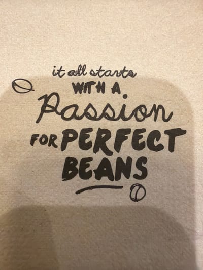The image shows a close-up of a beige, textured surface with black text. The text reads "it all starts WITH A Passion FOR PERFECT BEANS". The letters are in a mix of cursive and print styles, and some words are larger than others. There is an illustration of a coffee bean to the left and right of the text. The location in the city of Mole Valley, United Kingdom is not apparent from the image.