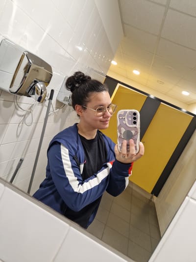 A young woman is taking a mirror selfie in a public restroom. She is wearing glasses, a black t-shirt, and a blue jacket with white stripes. She has her hair pulled up in a bun. The restroom has white tiled walls, a silver hand dryer, and yellow stall doors visible in the background. She is holding her phone, which has a purple case with white cloud and heart designs, and a circular pop socket. The image appears to be taken from a low angle, looking up at the woman. The specific location of Bahovitsa, Bulgaria is not identifiable from the image itself.