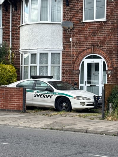 A photograph depicts a white "Palm Beach County Sheriff" vehicle parked on the sidewalk adjacent to a brick building. The building, identified by the number "85" on the door and the brickwork style, indicates that the location is the city of Leicester, United Kingdom. A satellite dish is fixed on the wall, and a window is visible. The vehicle is parked sideways, obstructing the pedestrian path. Details of the vehicle include the words "PALM BEACH COUNTY" and "SHERIFF" along with the number "206". A UK license plate "FX53" is seen on the front of the vehicle. The street is beside the pavement and a lawn is in front of the house.