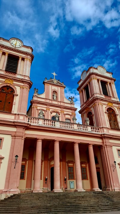 The image shows the exterior of the Göttweig Abbey, a Benedictine monastery located near Furth bei Göttweig, Austria. The photograph is taken from a low angle, looking up at the facade of the abbey church, which is characterized by its pink and cream-colored Baroque architecture.
The facade features two prominent bell towers flanking a central section topped with a cross. Both towers have clocks with Roman numerals and arched windows. The central part of the church has intricate sculptural details and a large balcony supported by columns. Below the balcony, a series of large, pink columns support the upper structure of the building. A wide, stone staircase leads up to the entrance.
The sky is a vibrant blue with scattered white clouds, suggesting it is daytime and the weather is fair. There are no people visible in the frame. The overall impression is one of grandeur and historical significance.