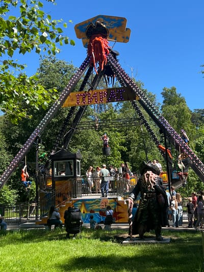 This is a medium shot, outdoor photograph taken on a sunny day at an amusement park in Zelenogorsk, Russia. The focal point of the image is a large, pirate-themed amusement ride with a pendulum-like structure. The ride is adorned with numerous colorful lights and features a prominent pirate skull and tentacles at the top. A banner with Cyrillic text and lights hangs across the structure.
In the foreground, a statue of a pirate with a long beard and tentacles for a face stands on a concrete base, facing the ride. To the left and right of the ride, various people are visible. Some are waiting in line, others are riding the attraction, and some are standing and observing. Children are also present, with one child sitting on a bench with an adult and another child walking near a stroller. The environment is lush and green, with grass in the foreground and mature trees in the background, indicating a park or outdoor recreational area. The sky is clear and blue.