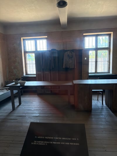 This image depicts the interior of a room, likely a former barrack, in Oświęcim, Poland. The room has wooden plank walls and flooring, with a large window featuring a grid of panes. Two striped shirts, characteristic of prisoner uniforms, are hanging on a wall. In the foreground, a dark informational plaque is visible with text in both Polish and English. The text reads: "TU MIESZKALI WIĘŻNIOWIE FUNKCYJNI OBSŁUGUJĄCY BLOK 11." and "THE HABITABLE ROOM FOR PRISONERS WHO WERE PERFORMING DUTIES IN BARRACK 11." This suggests the room served as living quarters for prisoners performing essential duties within Block 11, a significant part of the Auschwitz concentration camp complex. The overall impression is one of somberness and historical reflection, likely taken during daylight hours, given the natural light filtering through the window. The setting evokes a sense of the past, highlighting the harsh living conditions and the historical context of the location.