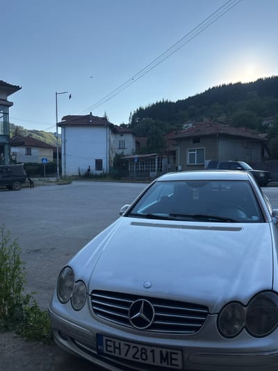 The image shows a silver Mercedes-Benz CLK-Class coupe parked on an asphalt surface, likely a street or parking area, in the town of Shipkovo, Bulgaria. The car is in the foreground, facing slightly to the left, with its front grille, headlights, and license plate visible. The license plate reads "EH 7281 ME". In the background, there are several houses with red tiled roofs, typical of Bulgarian architecture. A dark-colored SUV is parked further in the background. A street lamp is visible, as well as power lines crossing the sky. The sky is clear and blue, with the sun setting behind a forested hillside, casting a warm glow. A small bird is captured in flight against the sky. A black dog stands near the SUV. The overall scene depicts a quiet, rural or semi-rural setting in Bulgaria.