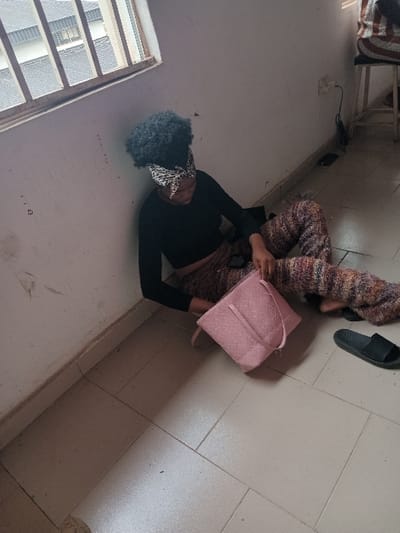 The image depicts an indoor scene, specified as being in OGBOKO, Nigeria. A female individual is seated on a light-colored tiled floor, leaning against a white wall in the foreground-left. She has dark, afro-textured hair secured with a white and black patterned headscarf. She is wearing a long-sleeved black top and multi-colored, textured wide-leg trousers. Her head is tilted downwards, and she is looking at a rectangular pink handbag with two top handles, which is positioned on the floor in front of her. Her hands are near or interacting with the handbag. Her left foot is bare, and a single black slide sandal rests on the floor to her right.
In the upper right background, a second individual is partially visible from the lower back and legs, seated on a wooden stool with a striped cushion (red, white, and brown pattern). The white wall behind the individuals shows some discoloration and dirt marks. A white power outlet with a black cable plugged in is visible on the wall to the right. A dark, rectangular object (possibly a mobile phone) lies on the floor near the cable. A window with horizontal metal security bars is present in the upper-left, showing the roof of an adjacent building under an overcast sky. The floor consists of light-colored, square tiles with visible grout lines.