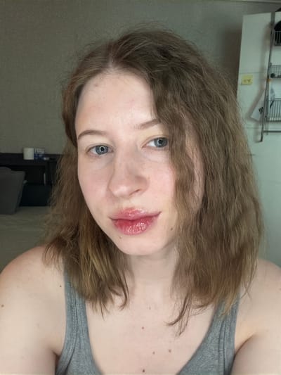 A young woman with light brown, wavy hair is pictured from the chest up, facing the camera. She has fair skin with some freckles, blue eyes, and is wearing a grey tank top. Her lips are glossy and appear to have a reddish-pink tint. The background is dimly lit and out of focus, showing a wall and a shelf with some items. The overall setting suggests an indoor residential environment. It is not possible to determine the exact location within Dongducheon-si, South Korea, from the visual information provided.