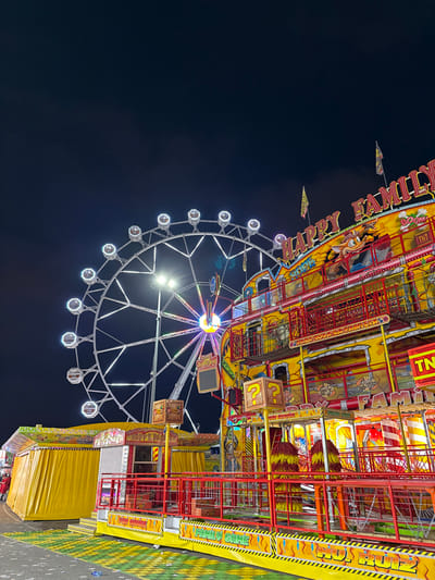 This image depicts a nighttime scene at a fairground in Polígon Industrial De Son Castello, Spain. The primary subjects are a large Ferris wheel and a multi-story amusement ride.
The Ferris wheel, positioned on the left, is white-framed with numerous enclosed gondolas, each illuminated by white lights. Its central hub displays a sequence of colored lights. The sky above is dark and cloudless.
To the right, a brightly illuminated, multi-level carnival attraction dominates the midground. Its primary colors are yellow and red, with black accents. The ride's top displays the words "HAPPY FAMILY" in large, stylized red and yellow letters, flanked by small flags. Decorative elements include cartoonish figures, text such as "BOOM," "TNT," "SHOOT GAME," "STOP GAME," and yellow blocks with red question marks. Red railings and transparent barriers define the levels. The lower portion features a sign reading "FAMILY GAME" and contains brush-like yellow and red elements.
To the far left, a smaller, single-story yellow booth with a canopy and a service window is visible, bearing text that appears to read "HAPPY FUN."
The ground directly in front of the rides is paved, covered in sections by a green and yellow patterned mat, and exhibits scattered debris or litter. No discernible individuals or specific actions are clearly visible within the frame. The overall environment is characterized by artificial light from the amusement rides against a dark sky.