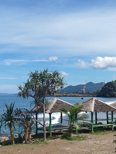 The image captures a coastal scene in Naga Umbang, Indonesia. The foreground features a dirt ground area with sparse vegetation, including small palm trees and bushes. Several thatched-roof pavilions with green wooden frames are positioned near the shoreline. The ocean is visible beyond the pavilions, with waves breaking along the rocky coast. In the background, a breakwater structure protects a harbor area, where large cranes are visible. A mountain range forms the backdrop under a partly cloudy sky.