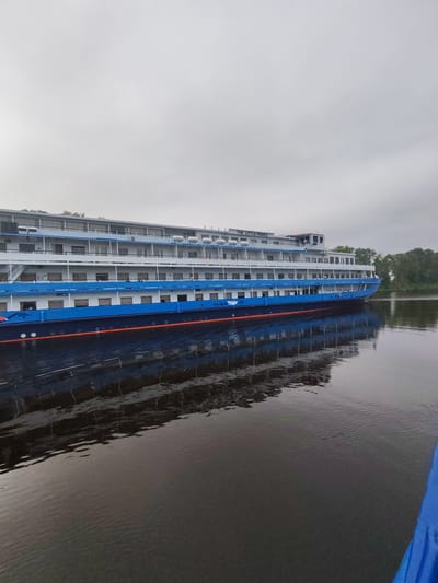 A wide, horizontal, eye-level shot captures a large, multi-deck passenger ship docked on a calm body of water. The ship, painted in shades of blue and white with an orange waterline, is the primary subject. The word "PRINCE" is visible in large, dark letters on the top deck, and "THE LITTLE" is positioned just above it, suggesting the ship's name is "THE LITTLE PRINCE." The ship has many windows and balconies visible across its decks. The water in the foreground is dark and reflects the ship, creating a distorted mirror image. In the background, a dense line of trees suggests a shoreline. The sky is overcast and a pale grey. The image appears to be taken from another watercraft, as a bright blue object, possibly part of a boat, is visible in the bottom right corner. No people are clearly visible on the ship or in the water. The overall impression is one of a quiet, overcast day.