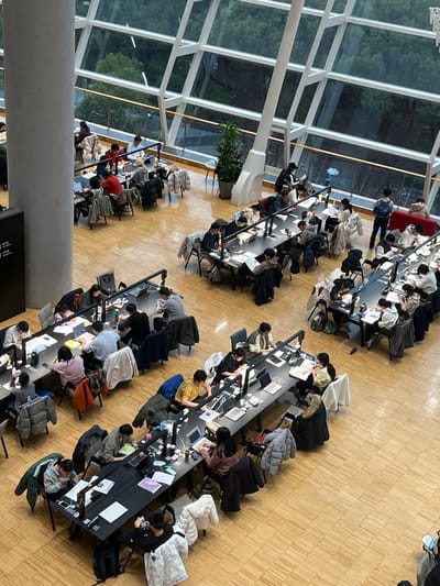 The image depicts a large, modern indoor study or work area within a building located in the city of Yanqiao, China. The space features a light-colored wooden plank floor and a high ceiling. A dominant architectural element is the expansive, multi-paneled glass wall on the right, framed by white angular structures, offering an exterior view of green trees and an overcast sky. A tall, light gray cylindrical pillar is visible on the left side of the frame.
Numerous long, dark rectangular tables are arranged in organized rows throughout the area. Each table is equipped with multiple integrated desk lamps providing localized illumination. Approximately 50 to 60 individuals, predominantly young adults of East Asian descent, occupy seats at these tables. Most are engaged in solitary activities such as studying, working on laptops (including several silver-colored models resembling MacBooks and other darker notebooks), reading physical books and papers, writing, or using mobile phones. Personal items like water bottles, coffee cups, and backpacks are present on the tables and floors. Many individuals are dressed in casual winter attire, with jackets and coats often draped over their chairs. A red upholstered sofa is visible in the upper right section, near the glass wall. A potted green plant is placed near the glass wall, and another smaller green plant is visible near a support structure. The environment suggests a quiet, focused academic or collaborative work setting.