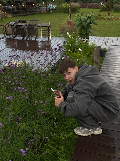 A young man with short brown hair, wearing a dark gray hooded sweatshirt and matching pants, crouches on a wet wooden deck. He is holding a white mobile phone in his hands, looking towards the camera with a surprised expression. Behind him, a patch of lush green grass is filled with blooming purple flowers and some white flowers. The wooden deck appears to be part of an outdoor seating area, with some chairs stacked on a table in the background, also wet from rain. The overall setting suggests a garden or patio area. The location is identified as Xuan Truong, Da Lat, Vietnam.