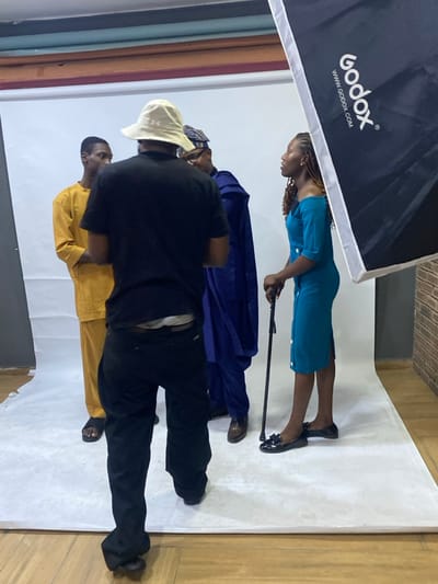 A group of people are standing in a photography studio. A woman in a blue dress holds a cane. A man in a blue robe stands next to her. Another man in a yellow robe is partially obscured by a man in a black shirt and black pants who is wearing a white hat. A large Godox softbox is visible in the upper left of the frame. There is a white backdrop and a row of fabrics to the side.