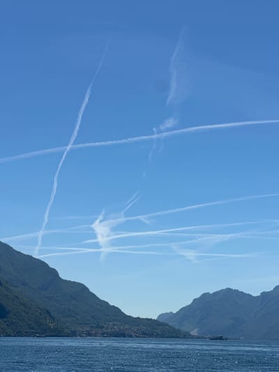 The image shows a bright blue sky with numerous contrails crisscrossing the expanse. In the foreground, a deep blue body of water, likely Lake Como, sparkles with sunlight. Along the far shore, a chain of lush green mountains rises, dotted with small towns and villages nestled at their bases. The mountains are densely covered with trees, and a ferry or boat is visible on the water in the distance. The presence of multiple contrails suggests a busy airspace. The location appears to be the town of San Siro, Italy, given the characteristic landscape of Lake Como.