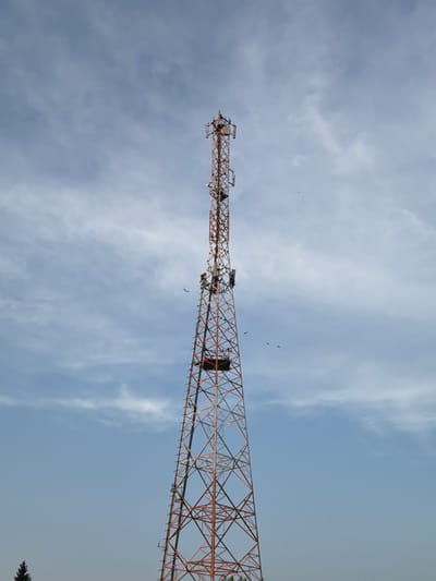 A tall, lattice cell tower with orange and silver supports rises against a pale blue sky with wispy white clouds. The tower is equipped with multiple antennas and appears to be operational. Several birds are visible in flight around the tower, with some perched on its structure. The base of the tower is not fully visible, but a small portion of a dark green evergreen tree is present in the lower left corner. The scene suggests an outdoor setting in or near the village of Vikulovo, Russia.
