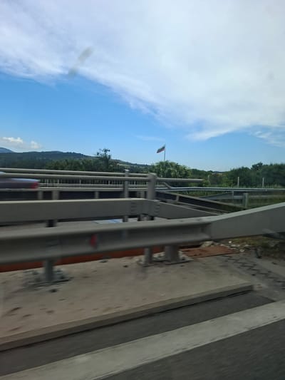 This media file shows a view from a moving vehicle, likely a car or bus, traveling on a highway in Eleshnitsa, Bulgaria. The scene is dominated by the sky and a roadside guardrail. The sky is a bright blue with wispy white clouds. The guardrail, made of metal, runs along the side of the road and is the primary foreground element.
Beyond the guardrail, a green, tree-covered landscape stretches into the distance, indicative of a rural or mountainous area. A flag, appearing to be the Bulgarian flag, is visible on a pole to the left, adding a specific location cue. The road surface appears to be asphalt, with some debris and construction materials scattered near the edge.
The time of day appears to be daytime, with clear skies and bright lighting, suggesting it is not dawn or dusk. The weather seems pleasant and sunny. The sense of motion is palpable, indicated by the slight blur in the image, suggesting the photograph was taken while the vehicle was in transit. There is no text visible in the image. The overall mood is one of travel and observation of the passing scenery.