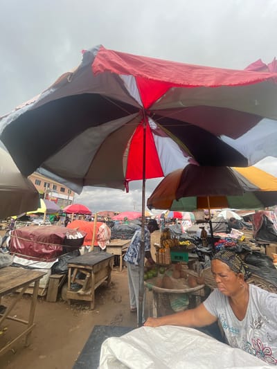 A bustling market scene in Ojodu, Nigeria. The foreground is dominated by a large, multicolored umbrella, its fabric faded and slightly tattered. A woman with a headscarf stands beside a table laden with coconuts, bananas, and other goods. The environment is crowded with stalls and people, with other umbrellas providing shade. Wooden tables and packed goods are scattered around. In the background, a building is partially visible. The ground appears uneven and possibly muddy. Overhead, a grey sky suggests overcast weather.