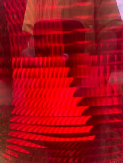 The image shows a blurry, predominantly red abstract pattern. A vague shape resembling a phone is visible, along with a person's reflection or presence behind the device. The red areas appear to be composed of numerous vertical lines with sharp edges, possibly reflecting a structured surface. The overall scene has a chaotic and unclear quality due to the blur.