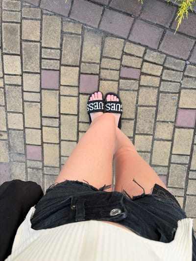 The image shows a close-up view of a person's legs and feet, as well as their black denim shorts and a white top. The person is wearing black slides with the word "GUESS" printed in white on the straps. Their toenails are painted pink. The background is a textured pavement made of interlocking bricks in shades of yellow, gray, and reddish-brown, with some greenery visible in the upper left corner. The lighting suggests it is daytime, and the overall scene appears to be outdoors on a paved walkway. The focus is on the clothing and footwear of the individual.