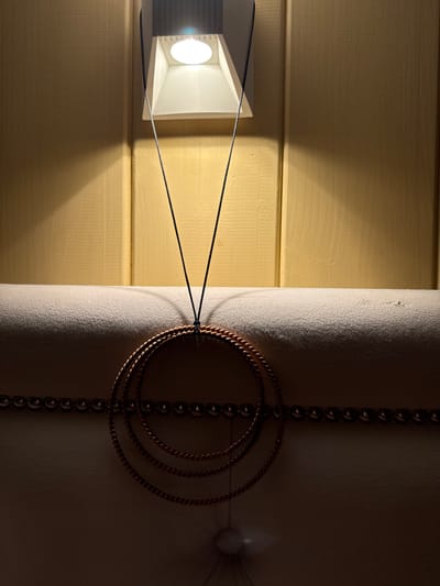 A close-up, low-angle shot captures a decorative element hanging in front of a light yellow, paneled wall. A white, recessed light fixture with a bright bulb is mounted at the top of the wall, casting a warm light and creating a distinct shadow on the wall to the left. A thin, dark string suspends a group of three concentric, twisted metal rings, appearing to be copper or brass. The rings are positioned over a padded headboard, which is upholstered in a light beige fabric and features a horizontal line of dark, rounded decorative studs along its top edge. The lighting creates a dramatic contrast between the illuminated upper wall and the darker, shadowed headboard area. There is no visible indication of the city of Liski, Russia, within the image itself.