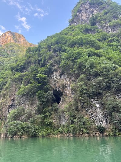 A vertically oriented photograph depicts a verdant mountain side meeting a body of water. The mountain is densely covered with trees and vegetation, clinging to the rocky terrain. A dark cave is visible near the water's edge, suggesting erosion or natural formation in the rock face. The water is a muted green color, with faint ripples disturbing its surface. A sliver of blue sky with scattered clouds is visible in the upper left corner, providing a contrast to the dominant green and gray hues of the landscape. The rugged texture of the rock is apparent, adding depth and dimension to the scene. Based on the prompt, the location is Đồng Văn Commune, Vietnam.
