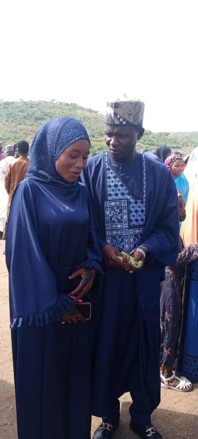 In Naraguta Hausawa, Nigeria, a man and a woman dressed in matching navy blue traditional attire stand together outdoors. The woman wears a hijab and holds a cell phone in her left hand, revealing henna designs on her hands. The man wears a patterned cap and a robe over a tunic with geometric designs. He holds a small green object in his hands. Other people are visible in the background. The environment appears to be a gathering or event with a slightly hilly landscape in the distance.